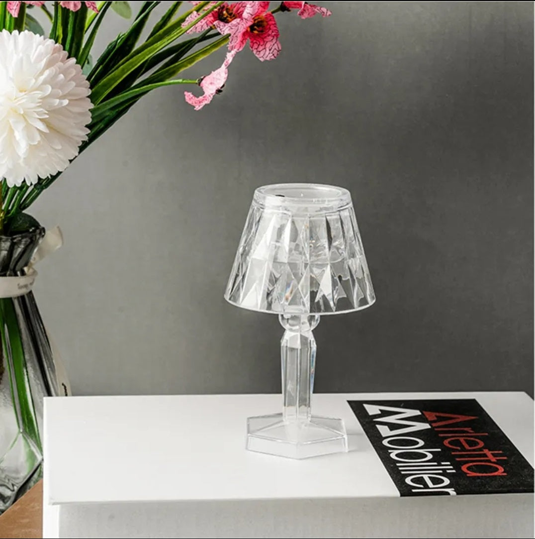 Clear table lamp on a white surface with flowers and a magazine in the background