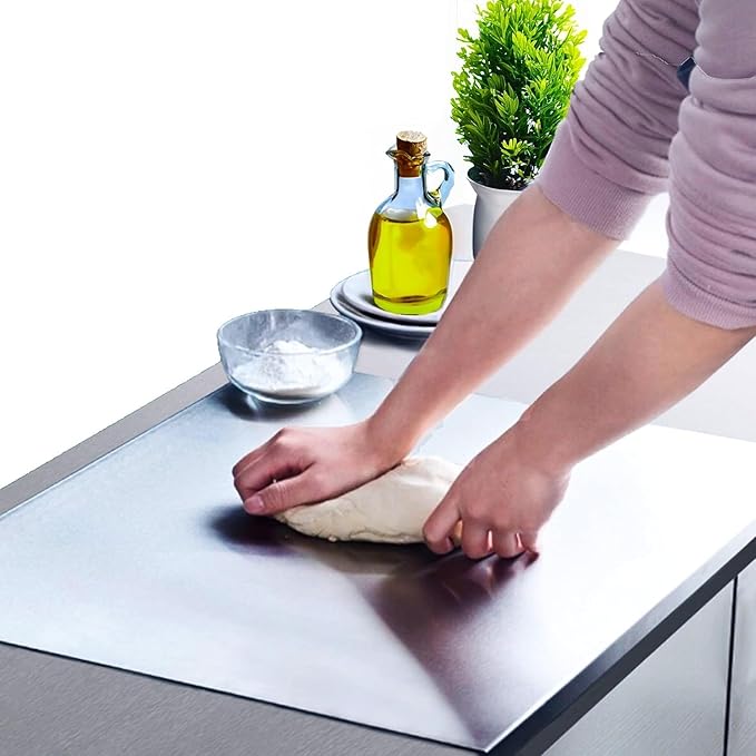 Person kneading dough on a stainless steel countertop with a bowl of flour and oil bottle in the background.