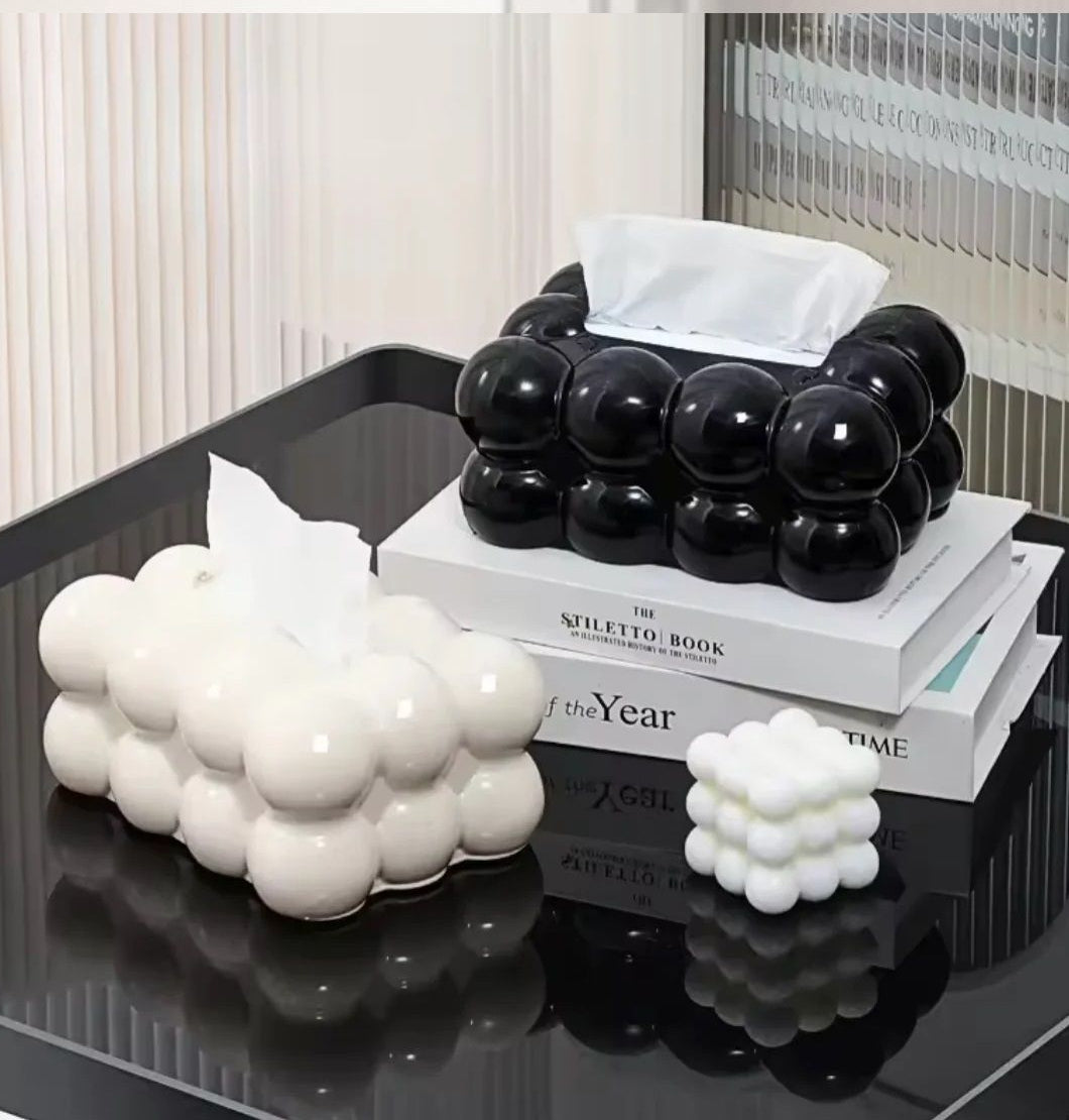 Black bubble-shaped tissue holder on a stack of books with white decorative items on a reflective surface.