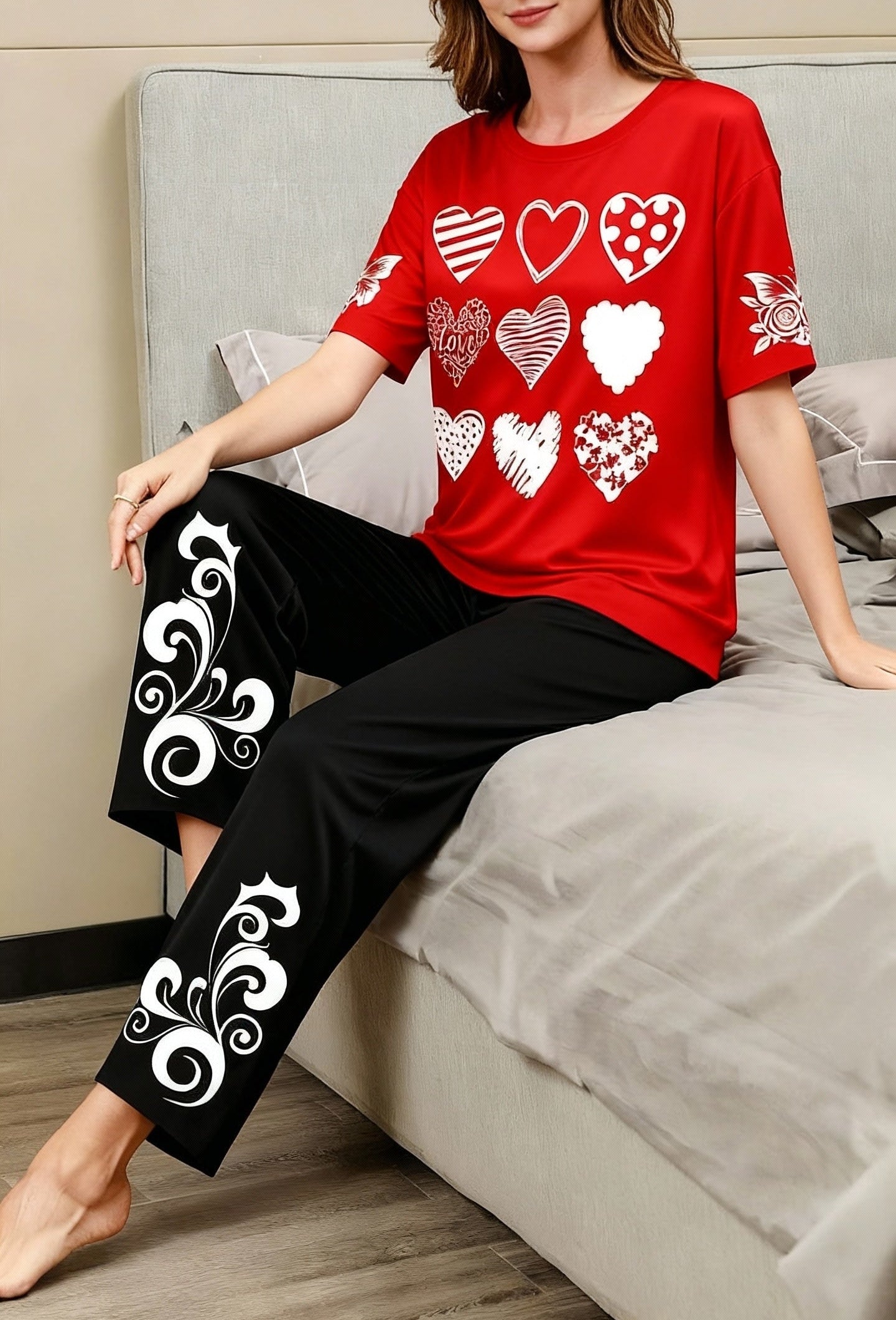 Woman wearing a red shirt with heart patterns and black pants with white designs, sitting on a bed.